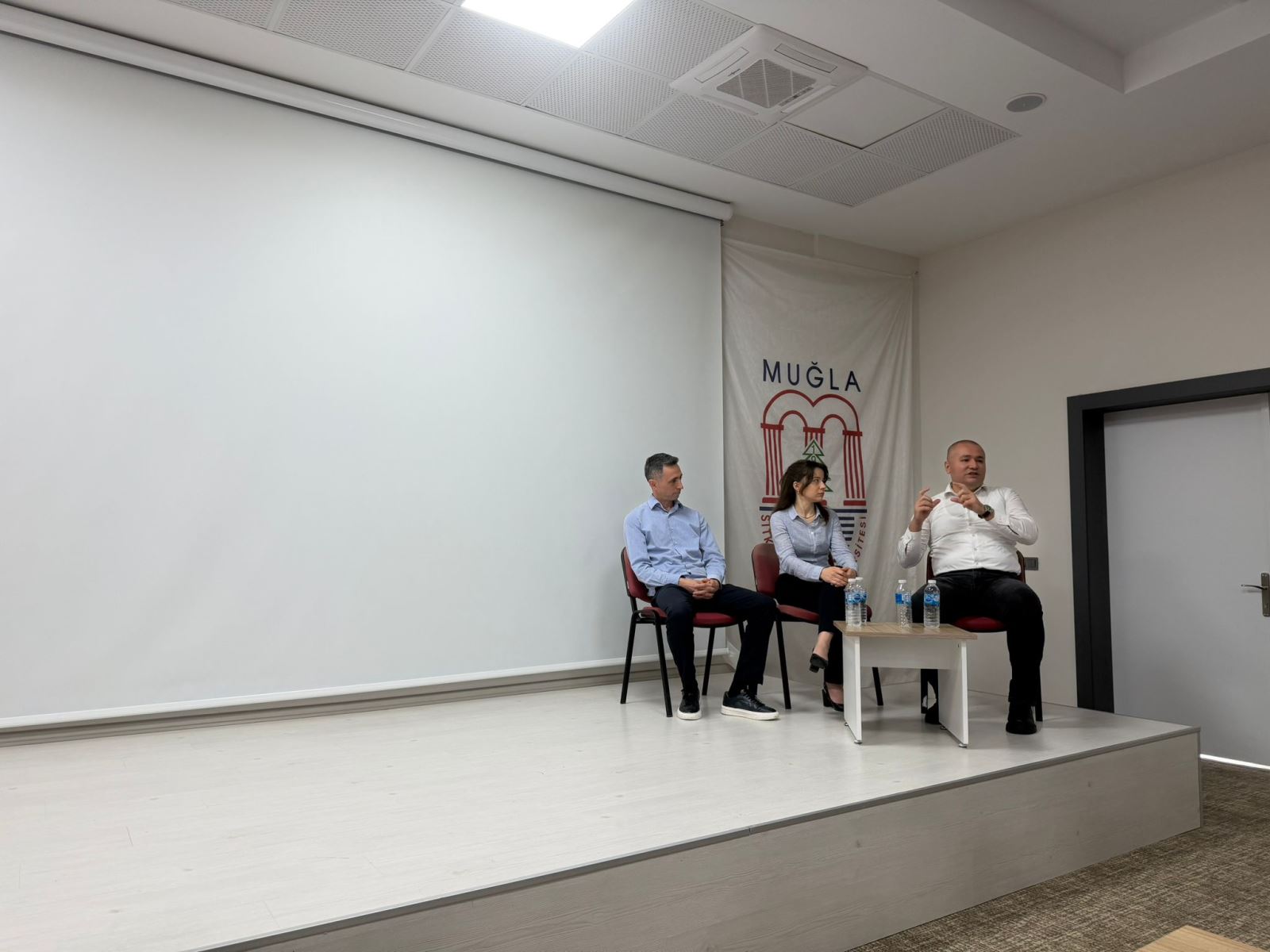 Sahne üzerindeki panel bölümünü yansıtan bu fotoğrafta, konuşmacıların bir arada olduğu samimi bir an yakalanmış. Konu detaylı bir şekilde anlatıldığı görülüyor, diğer konuşmacıların ise dikkatle dinlediği bu kare, interaktif ve tecrübe paylaşımına dayalı bir atmosfer sunuyor.